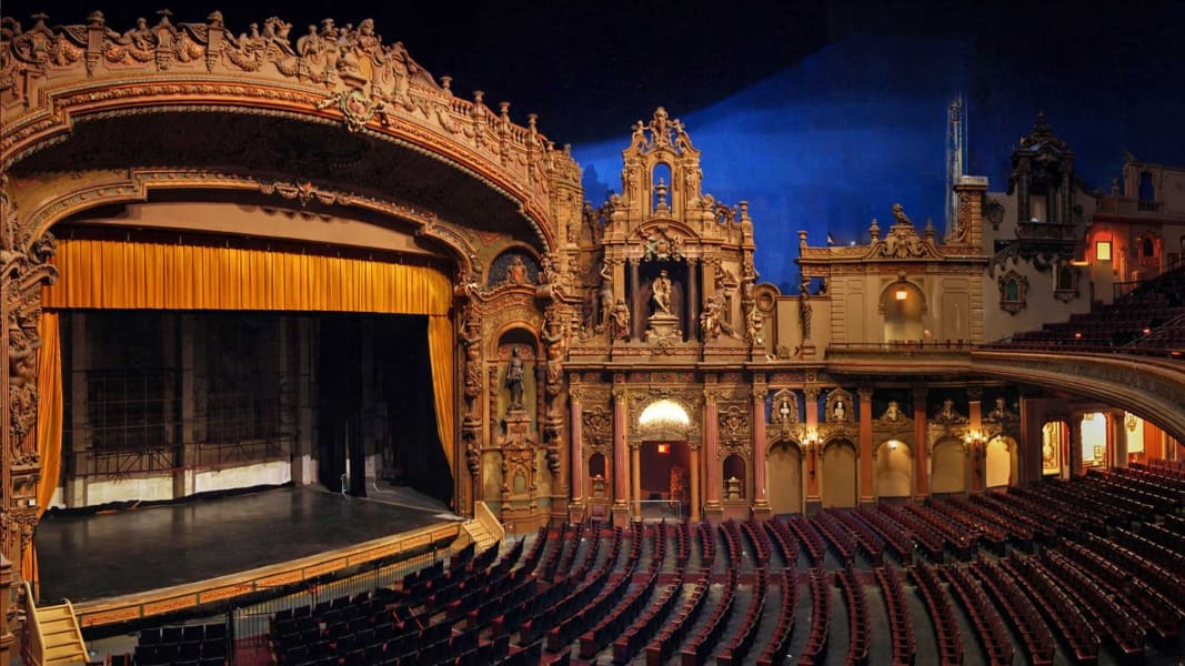 Ornate theater interior with red seats, golden decor, and a stage with a closed gold curtain under arch.
