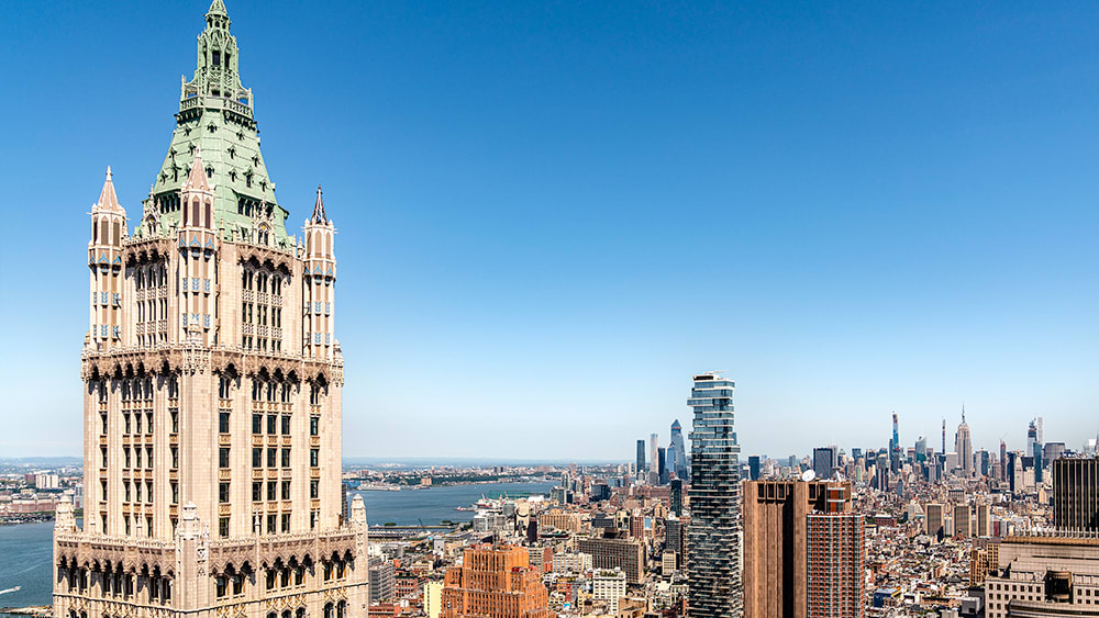 The crown of the Woolworth Building, an ornamental copper pyramid atop a heavily ornamented stone tower, and the cityscape of Manhattan looking north towards midtown and the Empire State Building.