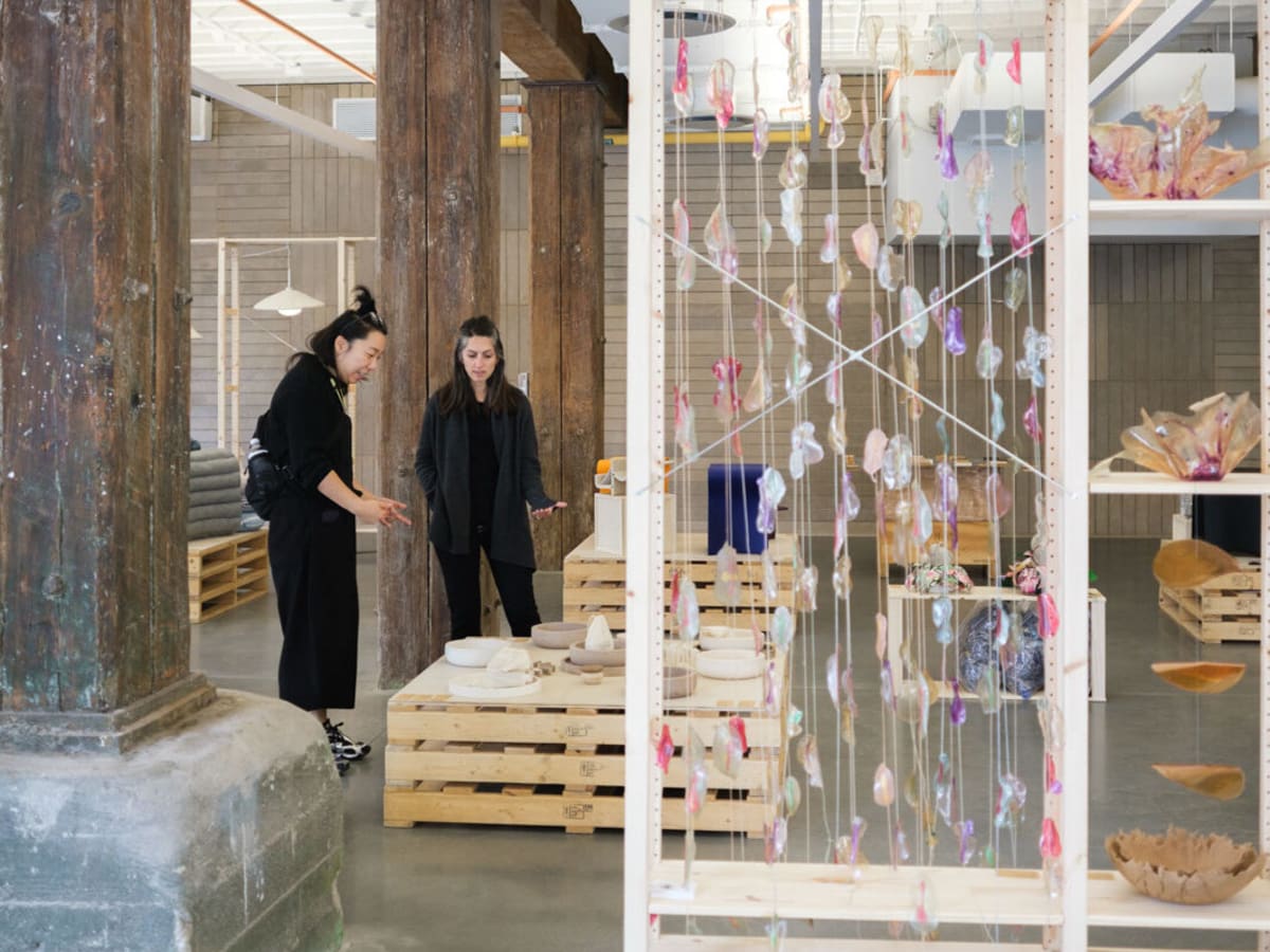 Two people stand in an art gallery with wooden columns, examining ceramic pieces on wooden pallets. Colorful glass or resin ornaments hang on strings in the foreground, partially obscuring shelves with more art objects.