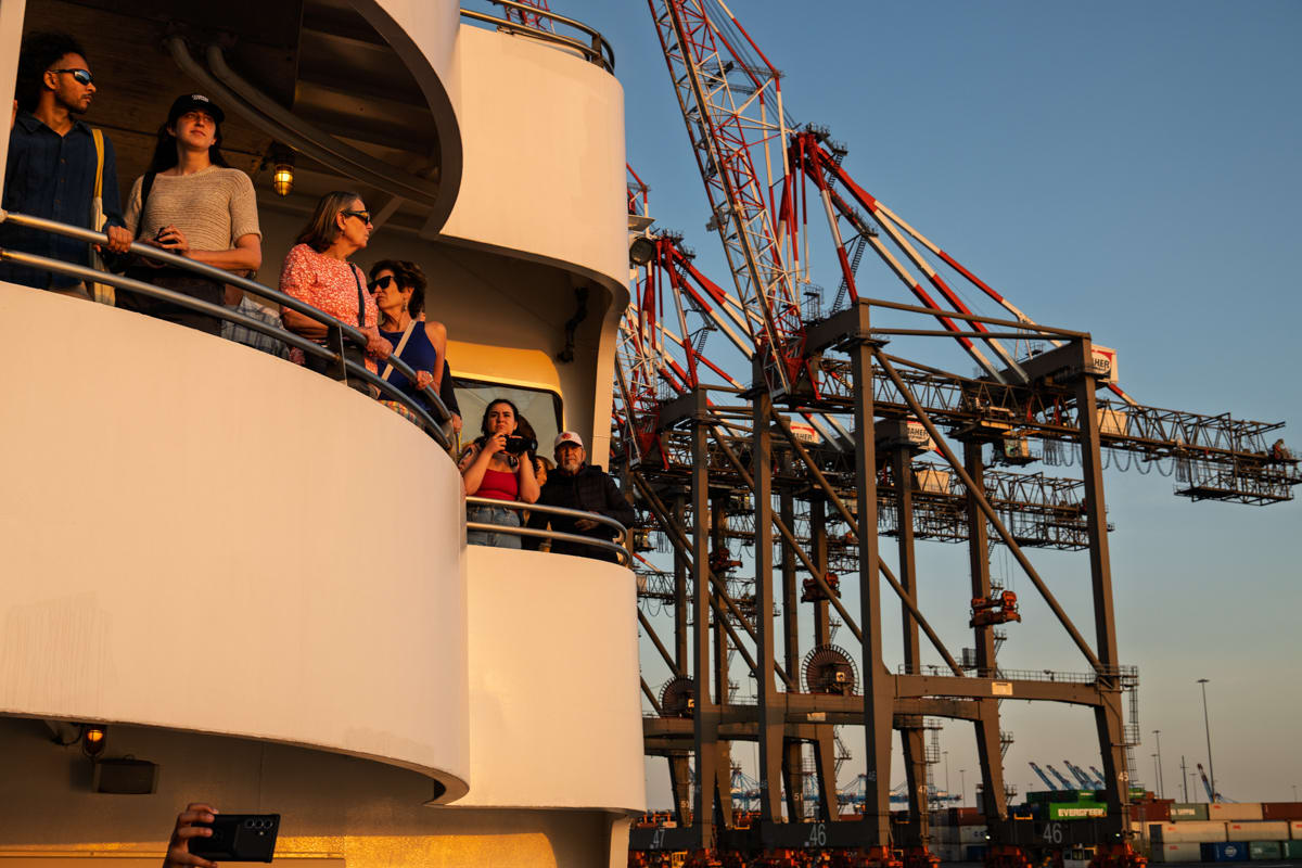 People on a ship deck look at cranes and stacked shipping containers at a port during sunset.