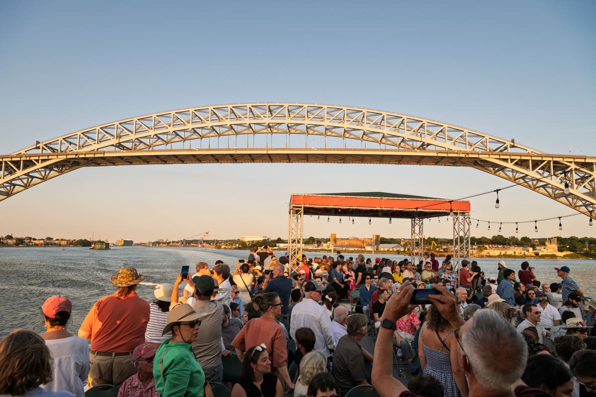 The top deck of a boat, crowded with people, that is siling through a waterway lined with gantry cranes. A large container ship is being loaded to the right, and in the distance, another container ship is sailing away.
