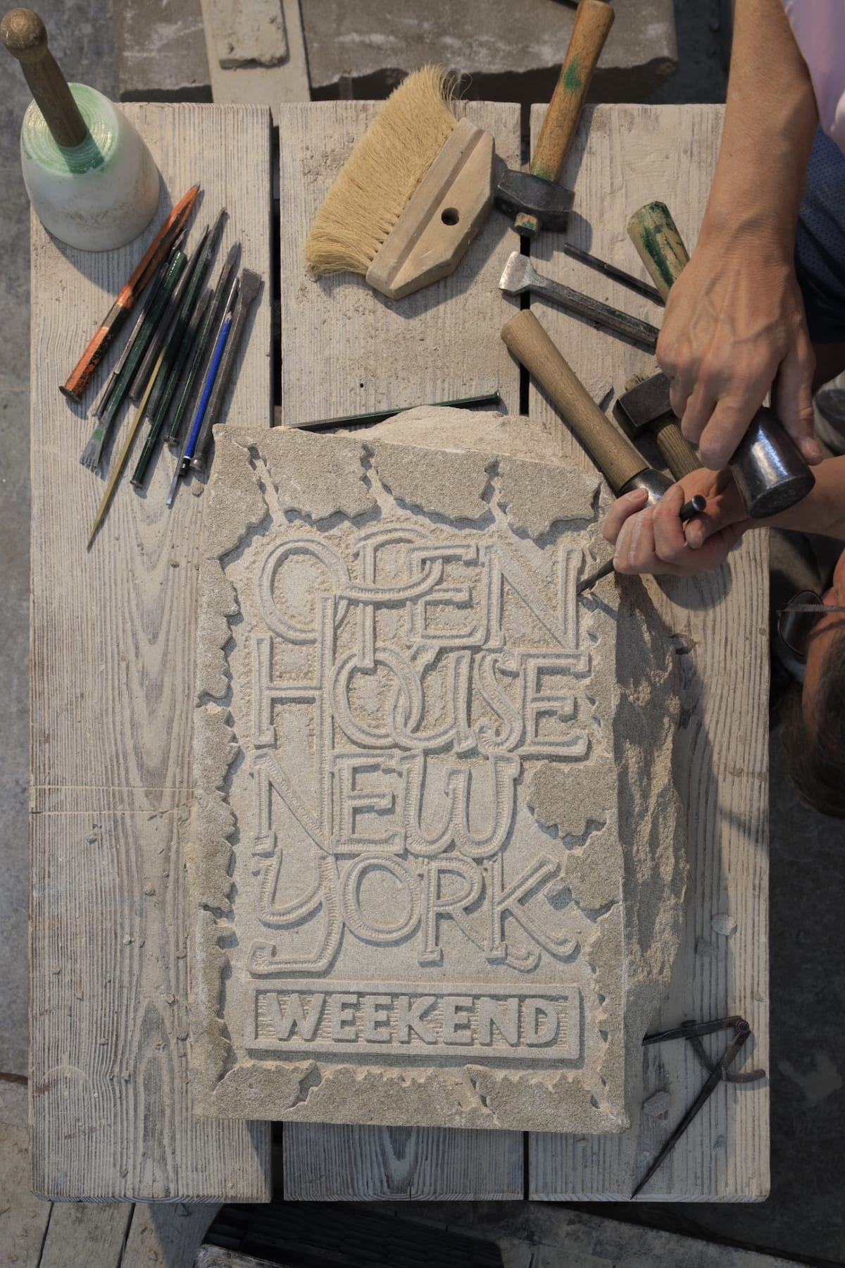 A person carves GLEN HOUSE NEW YORK WEEKEND into a stone slab on a wooden table with tools.