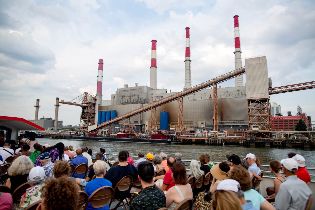 A large audience sits by a river, facing an industrial power plant with tall chimneys and red-striped stacks.
