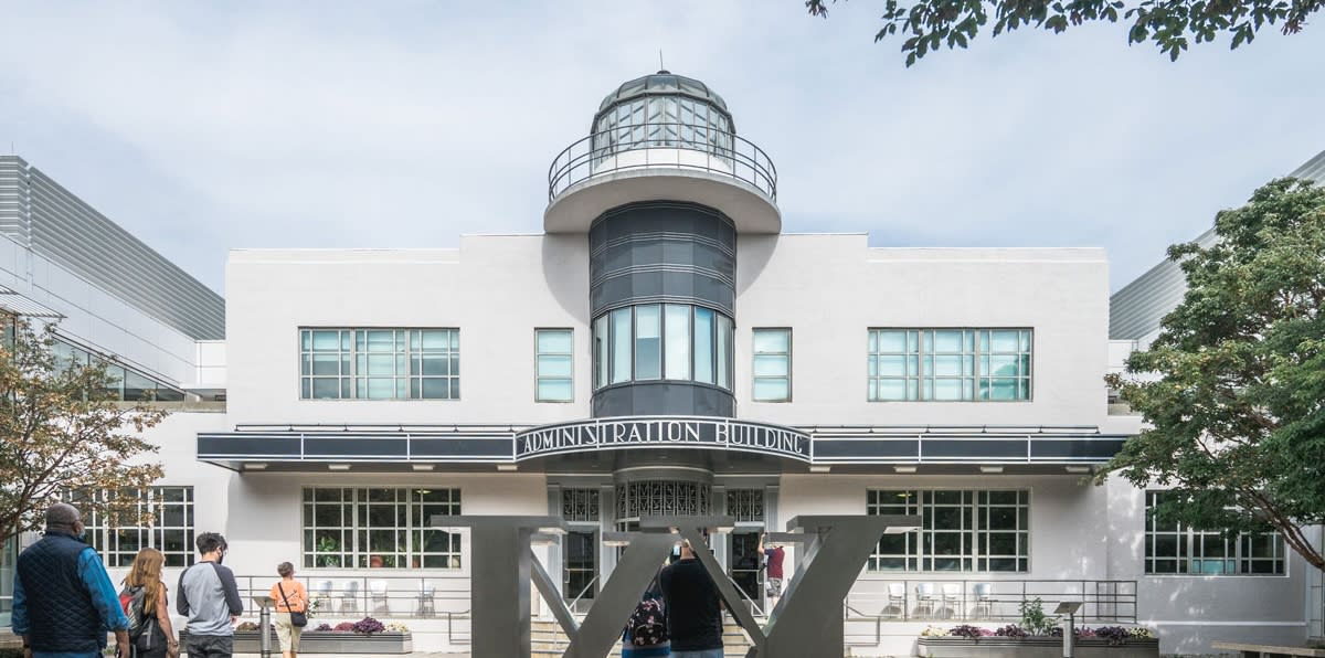 A square white building with a central grey tower and a steelcupola on top. An abstract steel sculpture sits outside and a group of people are walking toward the entrance.