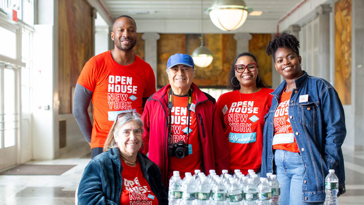 Five smiling people in Open House New York shirts stand behind a table with water bottles and papers.