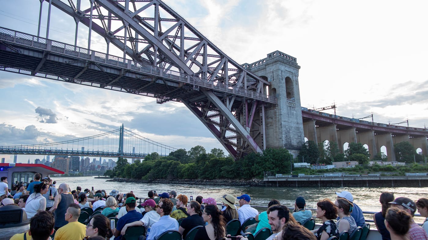 A boat with may people on the top deck passes beneath the Hell Gate Bridge.