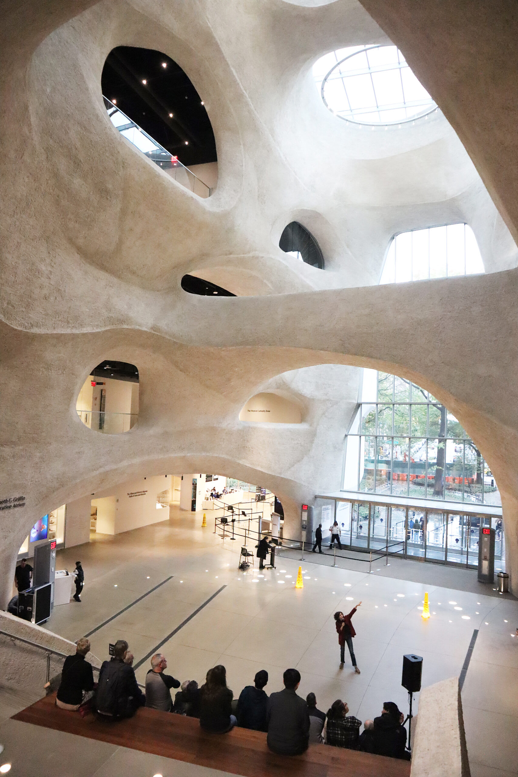 Spacious atrium with curved walls, skylights, and a presenter speaking to a seated audience on wooden steps.