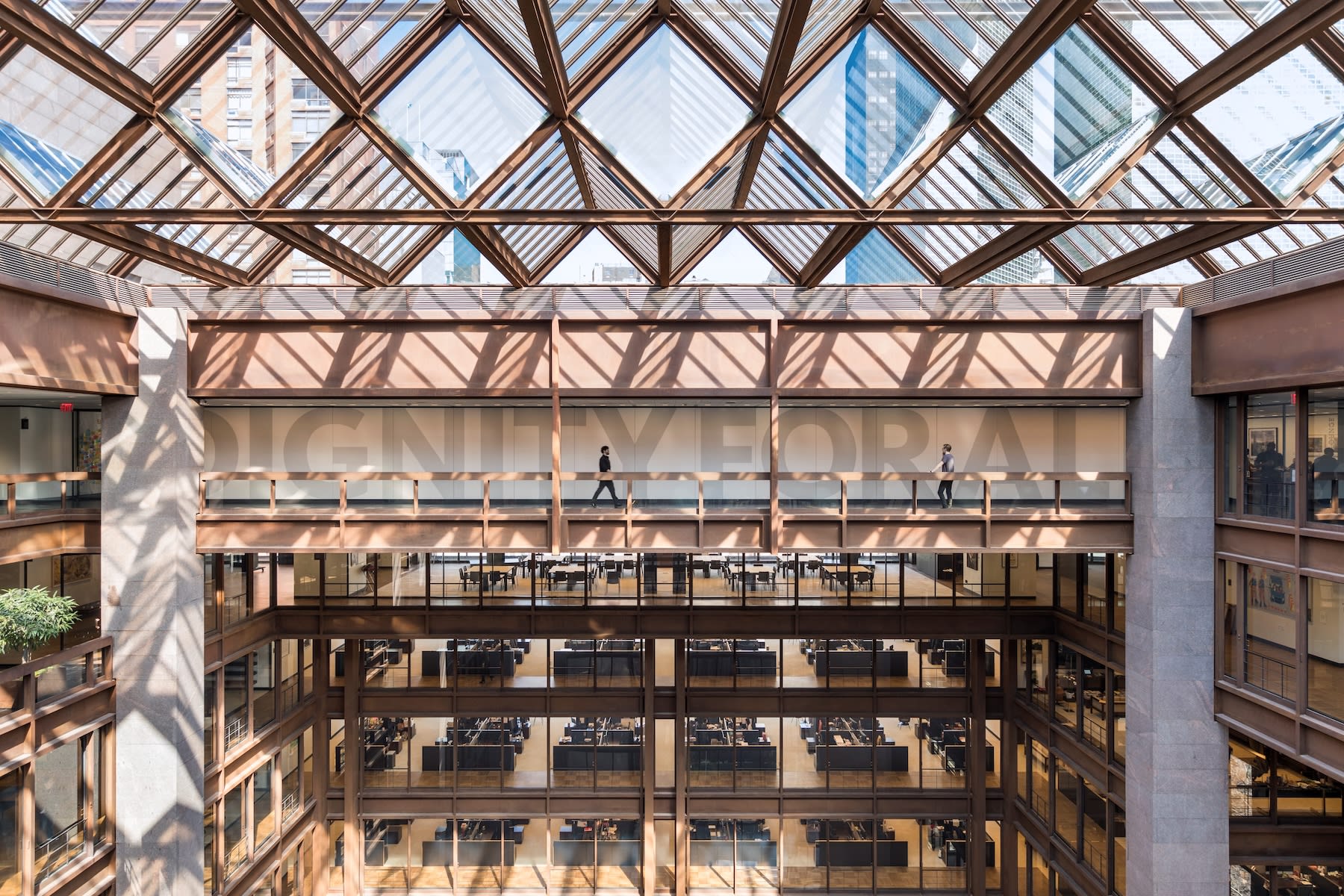 Spacious atrium with glass ceiling, people walking on upper bridge, and office desks below.