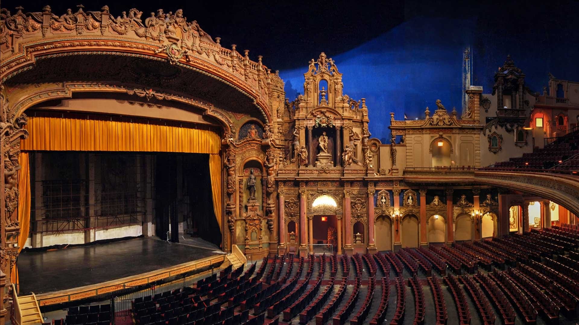 Ornate theater interior with red seats, golden decor, and a stage with a closed gold curtain under arch.