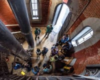 A group of people gathered inside a brick building with large black pipes, seen from above. Some stand while others sit along the wall near an open doorway and windows, suggesting a tour or meeting in an industrial space.