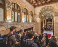 A group of people stand in an ornate, historic hallway with arched ceilings, detailed stonework, and wood accents, listening to a guide. Warm lighting highlights the architectural features.
