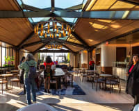 People stand and walk inside a modern, sunlit café with wooden ceilings, large windows, tables, chairs, and unique chandelier lighting. A few individuals are looking around or talking casually.