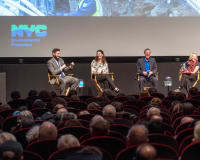 A large audience sits in a dark theater, watching a panel discussion on stage in front of a big screen.