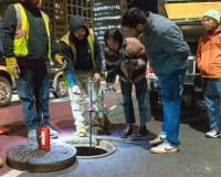 Four people look into an open manhole at night; two wear safety vests, others observe and take notes.