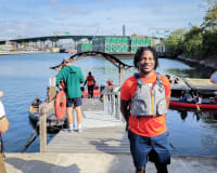 Smiling person in life vest stands on dock by water, with people and canoes in the background.
