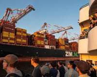 People watch a large container ship with cranes and stacked cargo moving past at sunset.