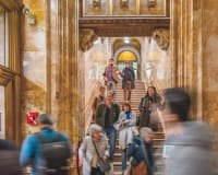A group of people stand in an ornate, historic hallway with arched ceilings, detailed stonework, and wood accents, listening to a guide. Warm lighting highlights the architectural features.