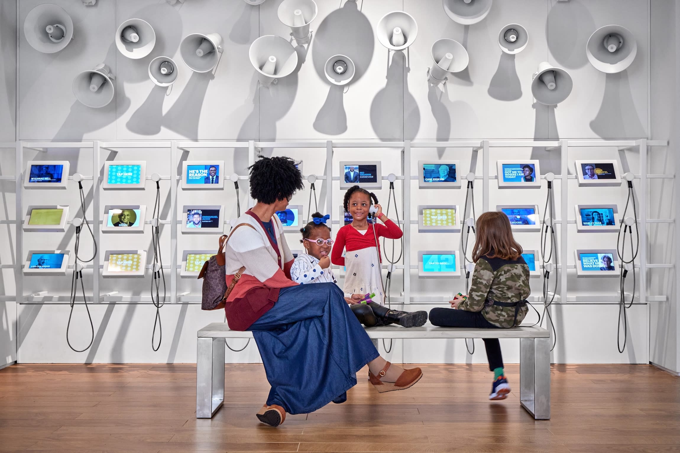 Children sit on a bench in a modern exhibit with tablets and wall-mounted speakers behind them.