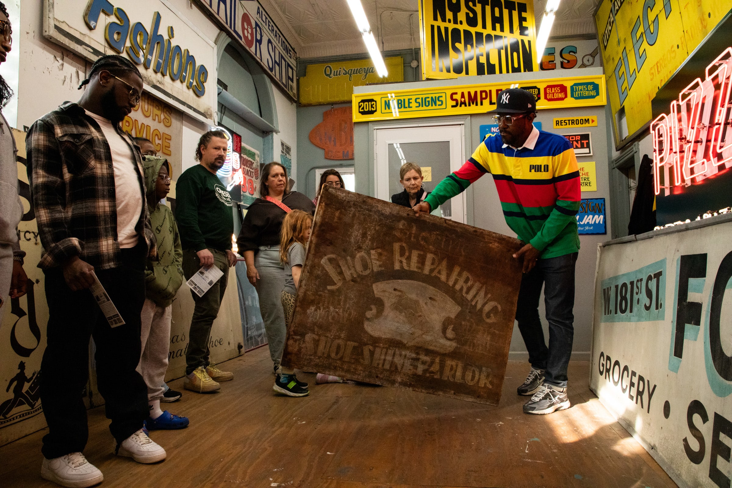 A group of people observe a man holding a large, weathered Shoe Repairing sign indoors.