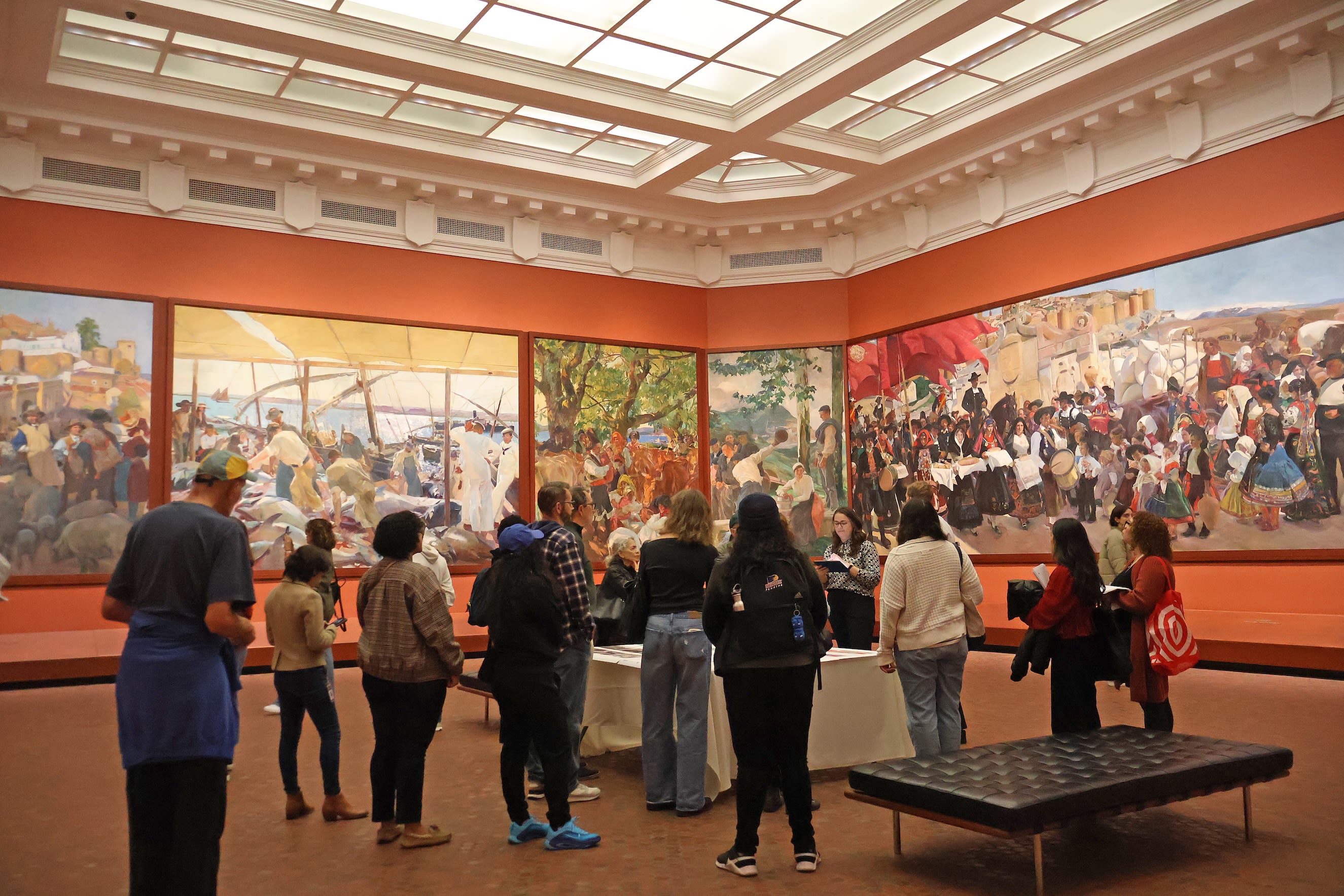 A group of people view murals and displays in a brightly lit museum gallery with orange walls.