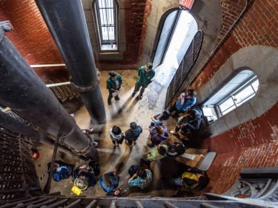 A group of people gathered inside a brick building with large black pipes, seen from above. Some stand while others sit along the wall near an open doorway and windows, suggesting a tour or meeting in an industrial space.