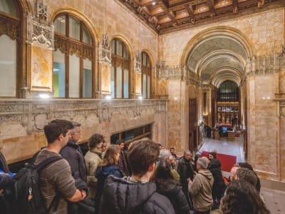 A group of people stand in an ornate, historic hallway with arched ceilings, detailed stonework, and wood accents, listening to a guide. Warm lighting highlights the architectural features.