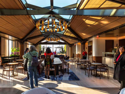 People stand and walk inside a modern, sunlit café with wooden ceilings, large windows, tables, chairs, and unique chandelier lighting. A few individuals are looking around or talking casually.