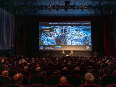 A large audience sits in a dark theater, watching a panel discussion on stage in front of a big screen displaying an aerial image of a construction site and the text “Water Talks: Inside DEP With Commissioner Aggarwala.”.