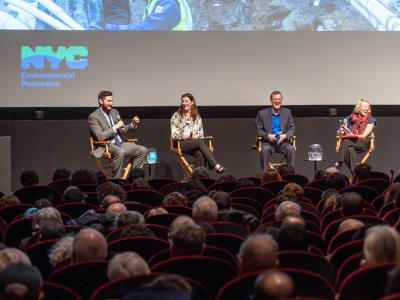 A large audience sits in a dark theater, watching a panel discussion on stage in front of a big screen.