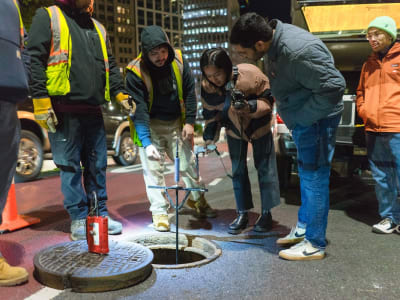 Four people look into an open manhole at night; two wear safety vests, others observe and take notes.