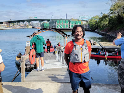 Smiling person in life vest stands on dock by water, with people and canoes in the background.