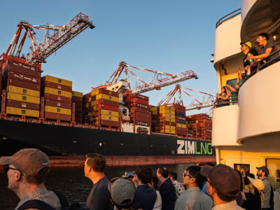 People watch a large container ship with cranes and stacked cargo moving past at sunset.