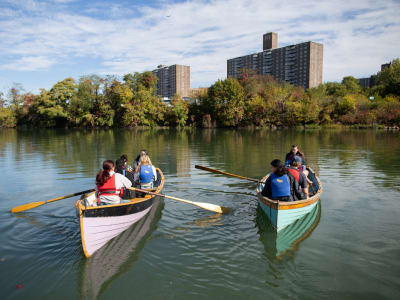 Two canoes with several people paddling on a calm river, surrounded by trees with autumn foliage. Tall apartment buildings are visible in the background under a partly cloudy sky.