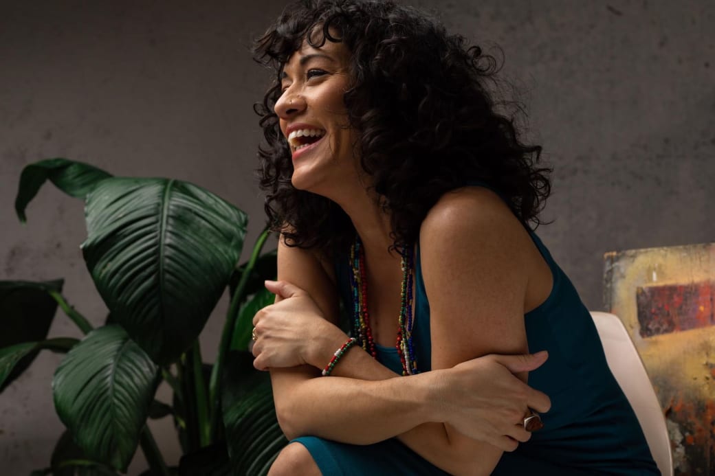 A woman smiling while sitting. There is a potted plant in the back