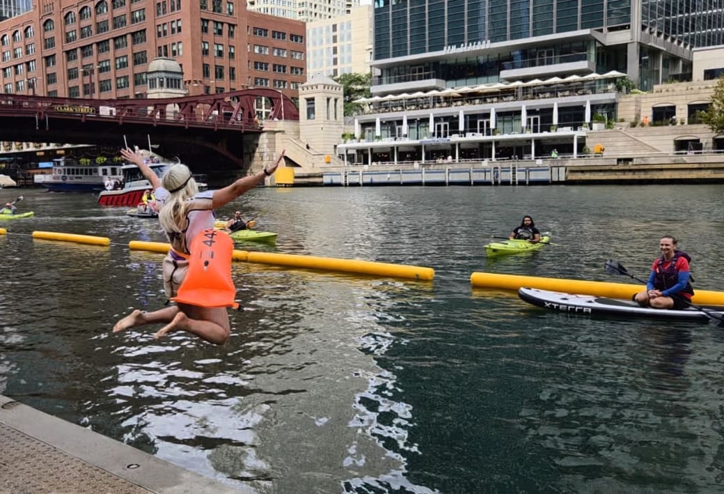 Emmi Mack jumping mid air into a greenish river. In the background there are people on canoes and a boat passing by. There is also a bridge and buildings in the background