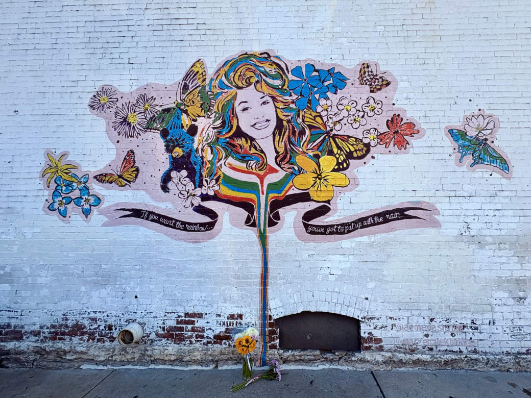 A mural of Dolly Parton on the side of a white brick wall. The mural has rainbow colors with butterflies and flowers. The words “If you want the rainbow, you've got to put up with the rain” is in a ribbon around the musician’s portrait.