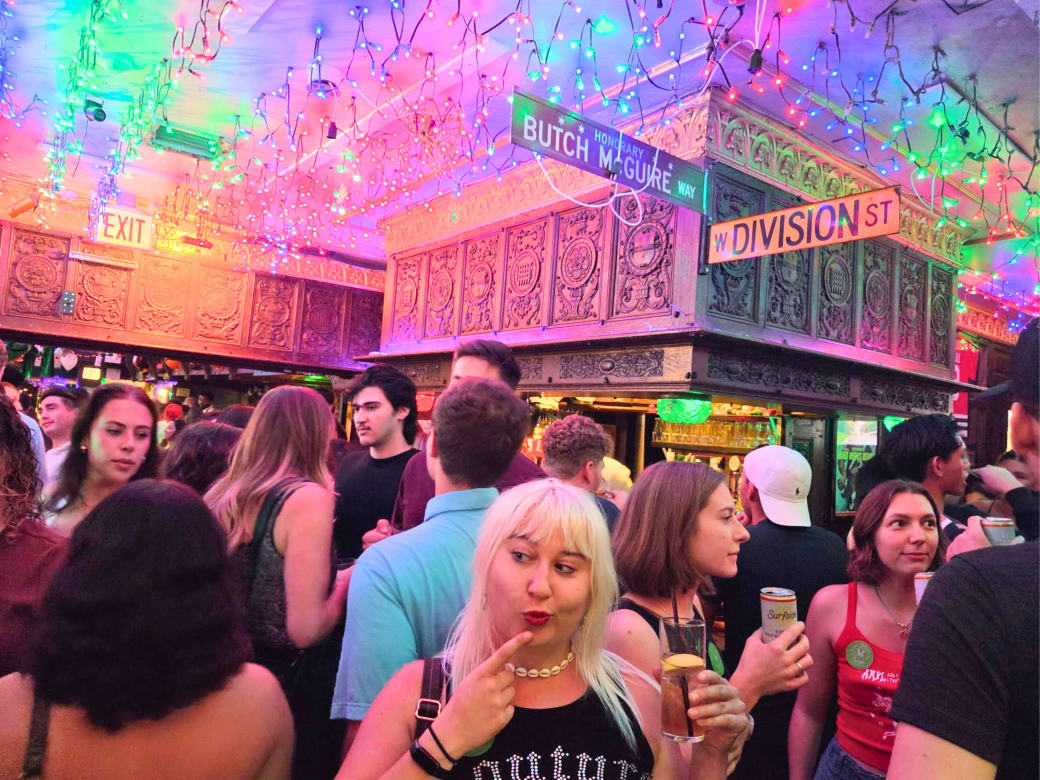 Emmi Mack in a crowded bar holding a drink posing with a look on her face as if she was deciding. She is surrounded by other people talking amongst themselves