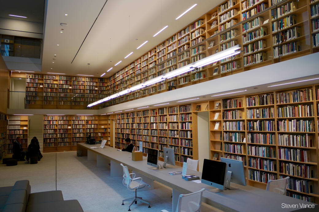 Two level room with floor to ceiling bookshelves filled with books. On the floor level is a long table has computers on it.
