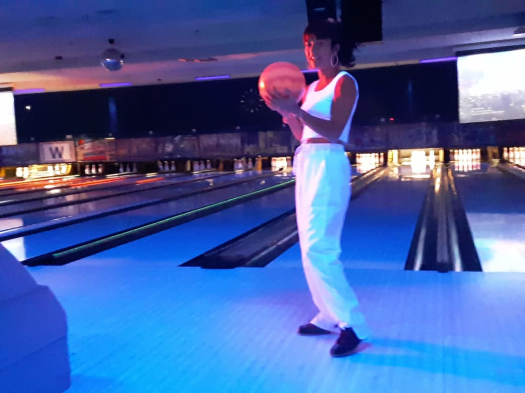 Woman lit by black lights stands in front of bowling alley lane holding ball and facing camera