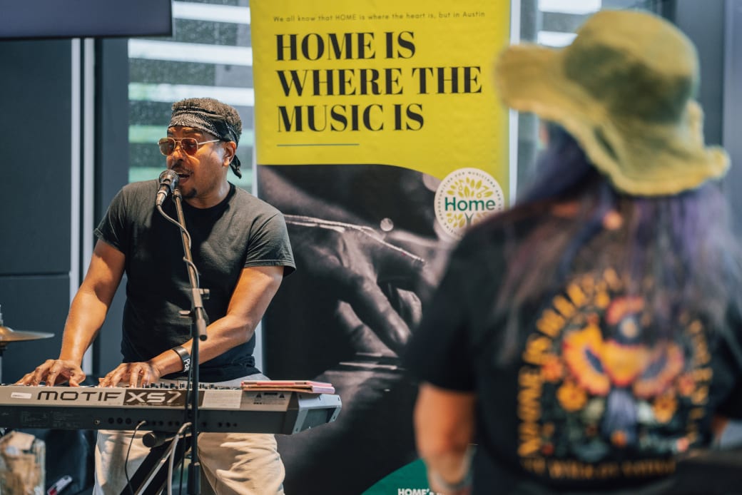 A person playing a piano and singing. A sign that says "home is where the music is" is behind them.
