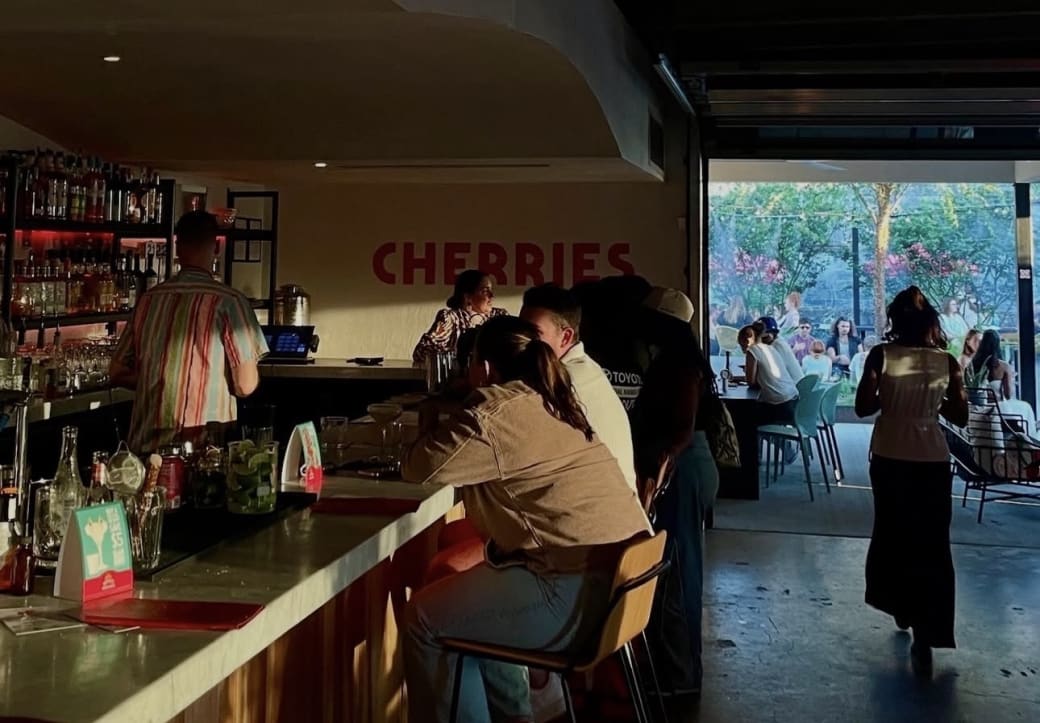 People gather at a bar with the word Cherries on one white wall, and a busy patio in the background.