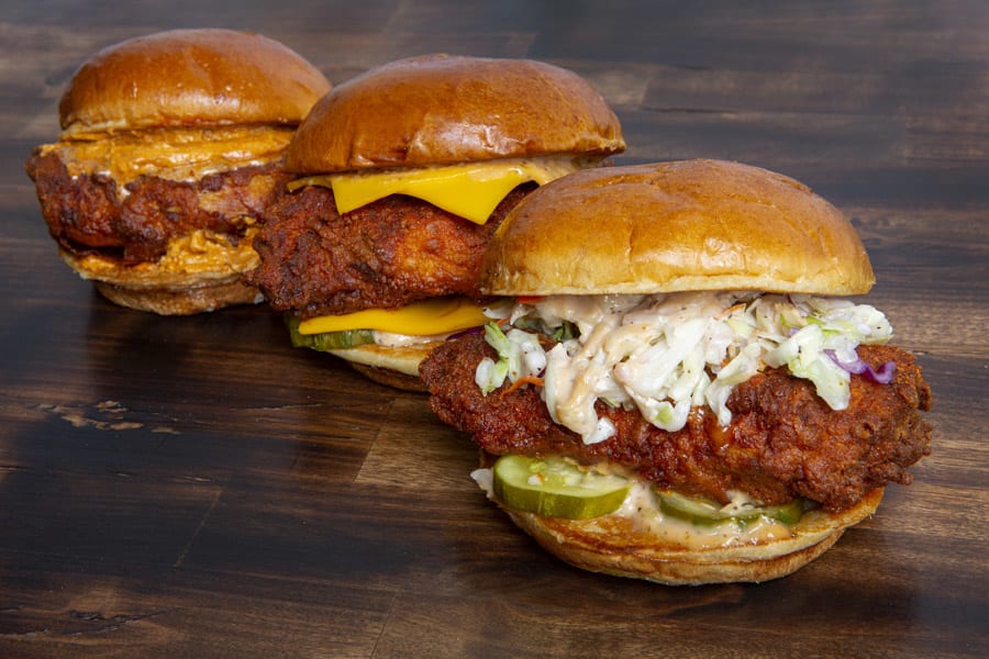 Three hot chicken sandwiches with various toppings lined up on a wooden surface