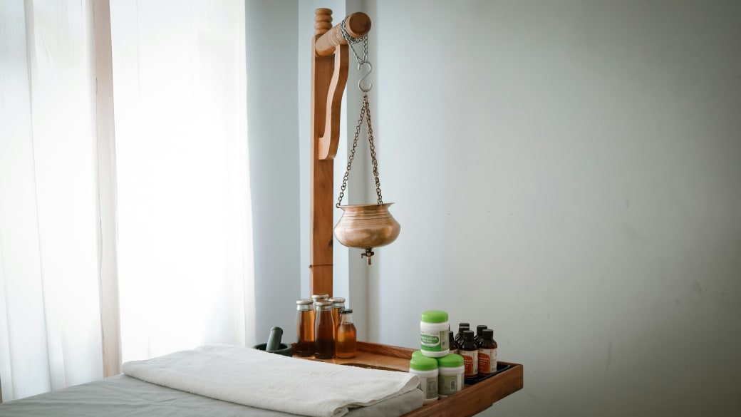 A wooden spa table with oils and a white towel in a white room. There's a bronze bowl hanging from a chain.