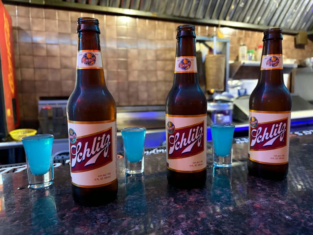 Three bottles of Shlitz with three shots of blue liquid on a bar top