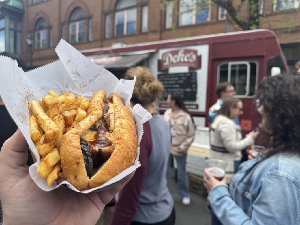A hand holds a BBQ sandwich and fries in front of Deke's BBQ food truck