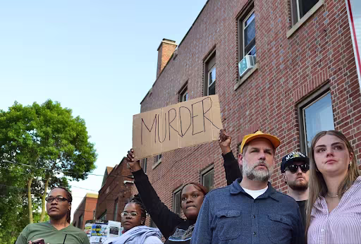 A group of protestors stand outside a brick building. One holds a sign that reads "MURDER."