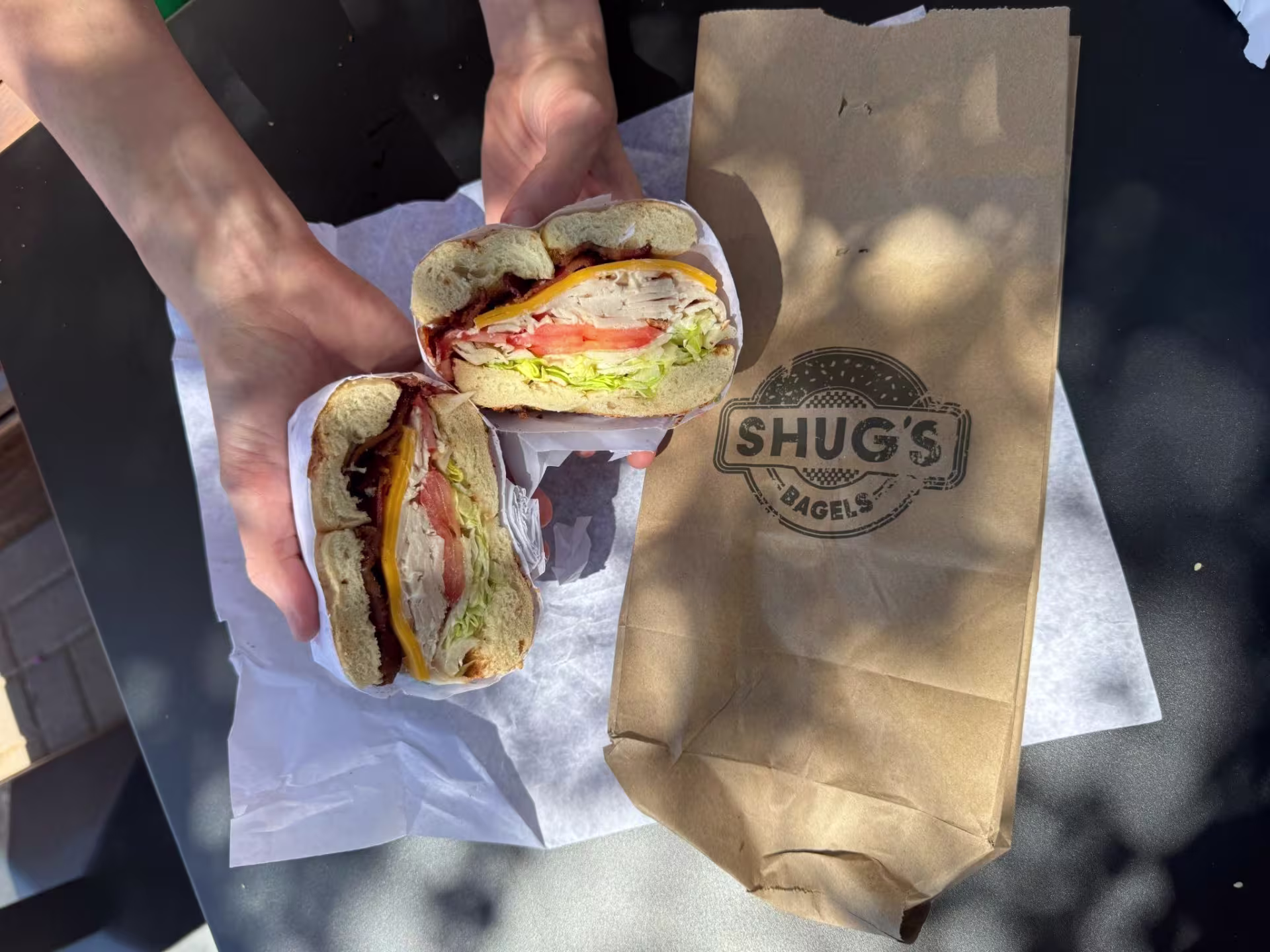 A person holding a bagel next to a brown bag that says "Shug's Bagels."