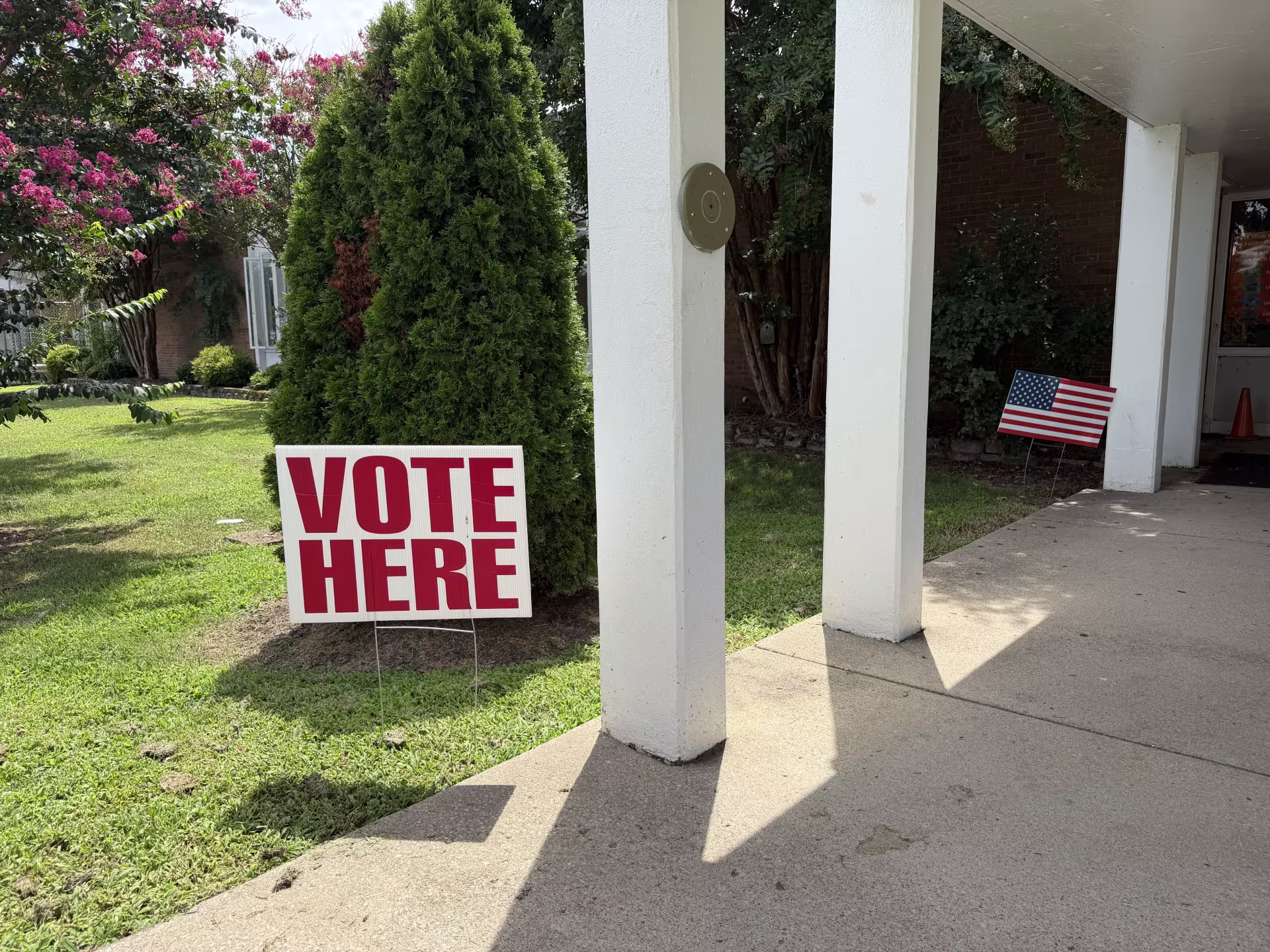 A white sign with VOTE HERE in red letters by a covered walkway, and in front of green shrubs. An American flag is in the background.