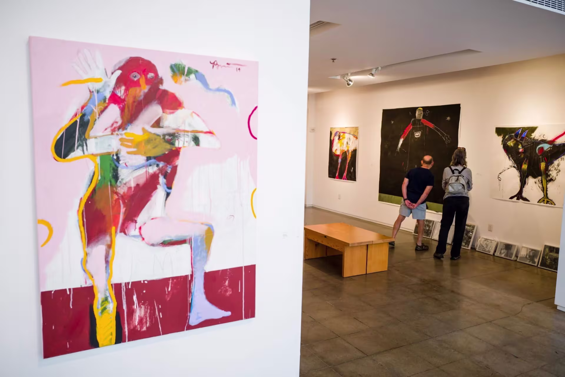 A colorful abstract painting of a person hangs on a wall to the left. In the background, two figures observe different paintings at the Froelick Gallery in Portland, Oregon.