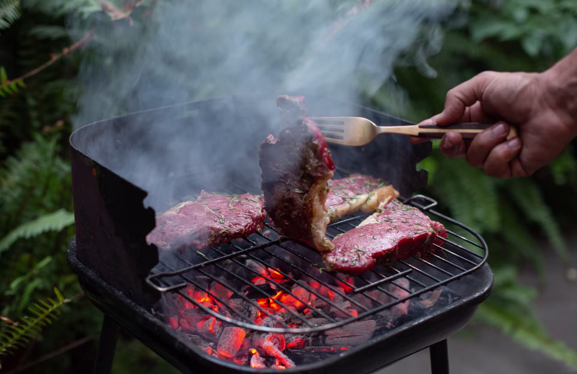 A small barbecue with two pieces of meat on it. A man is using a fork to lift one of the pieces of meat.